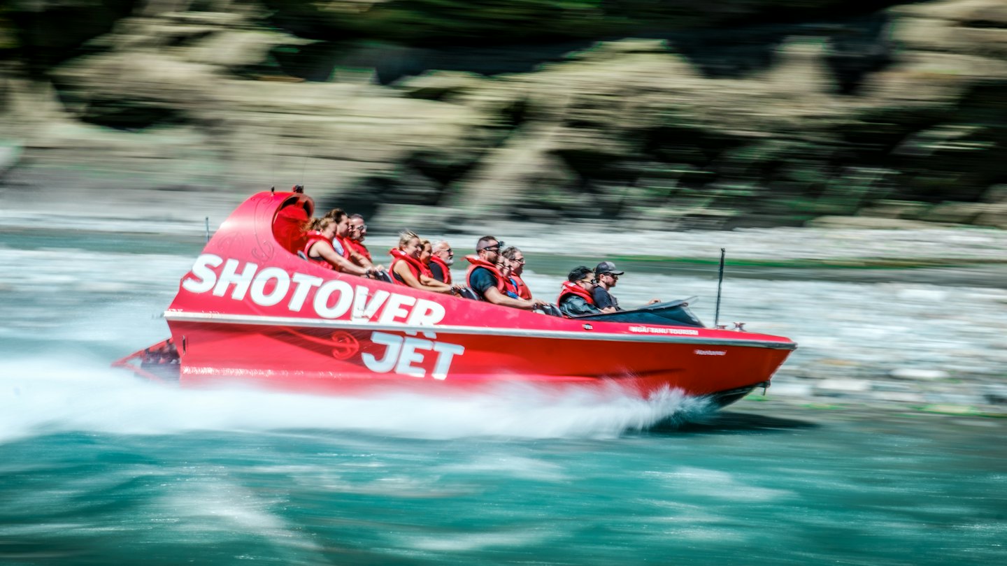 Red jet boat with "SHOTOVER JET" logo speeds through turquoise water.